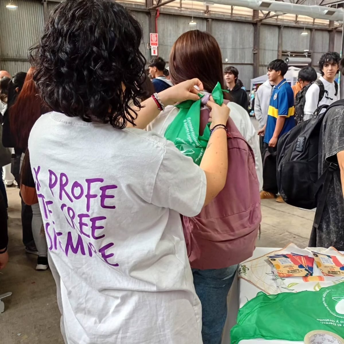 Integrante de la Red con remera violeta que dice «Tu profe te cree siempre» atando un pañuelo verde en la mochila de una estudiante, durante una feria